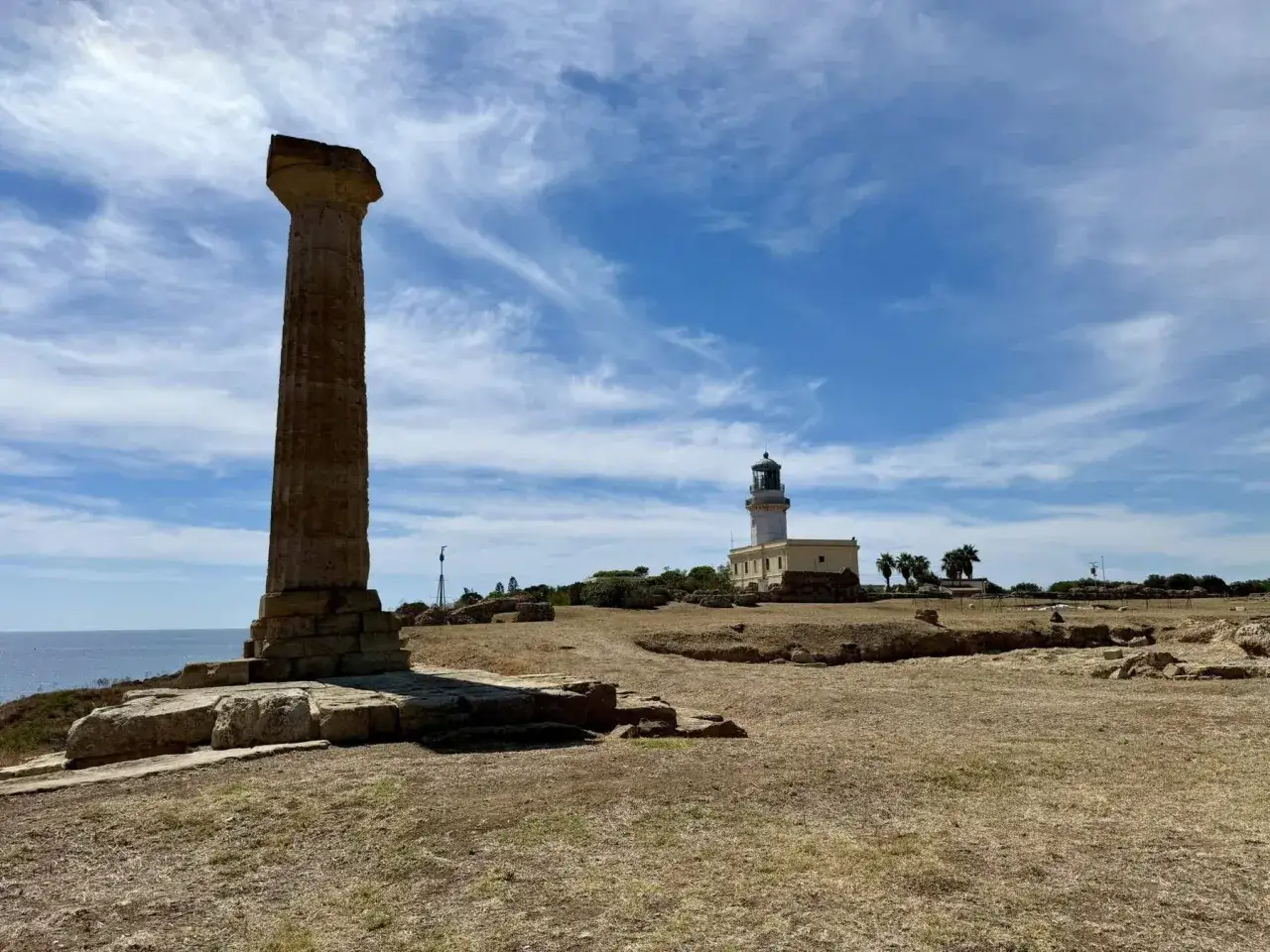 Antica colonna dorica e faro bianco, scorci di cosa vedere a Crotone, con il mare sullo sfondo e un cielo azzurro con nuvole bianche.