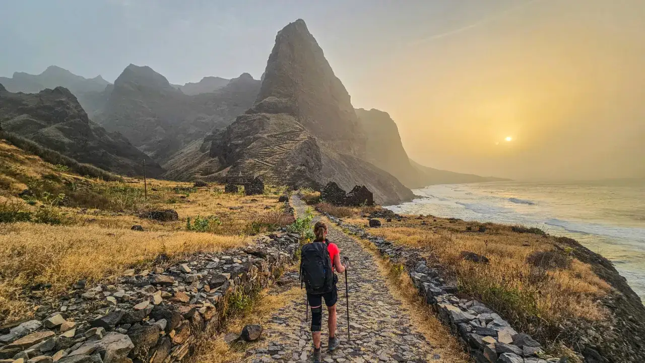 Escursionista su sentiero di pietra, tra rovine e montagne scoscese, con oceano e tramonto. Ecco cosa vedere a Capo Verde per un'avventura indimenticabile.