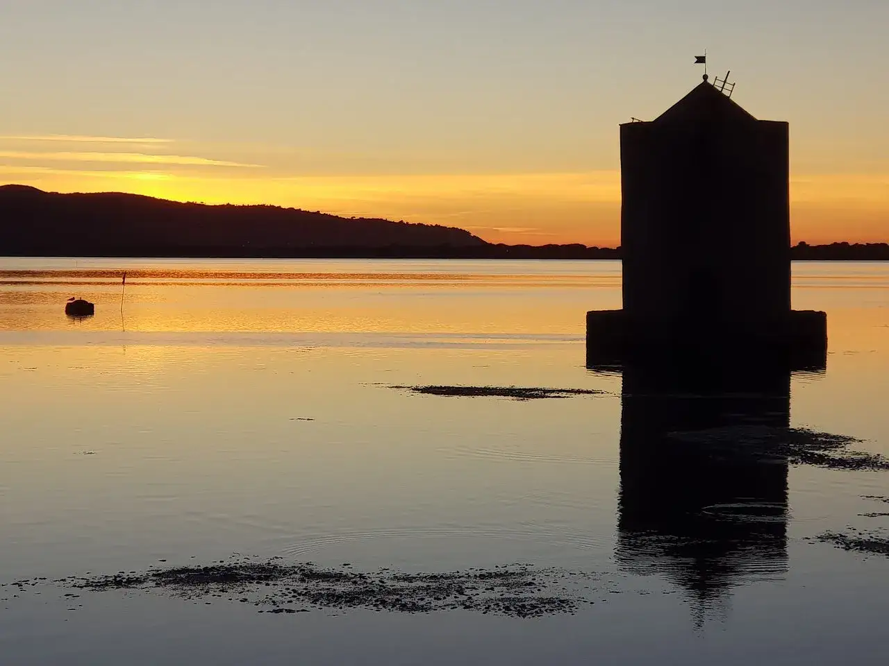 Torre di San Pancrazio a Orbetello, cosa vedere: silhouette di una torre sull'acqua al tramonto, con riflessi dorati e colline all'orizzonte.