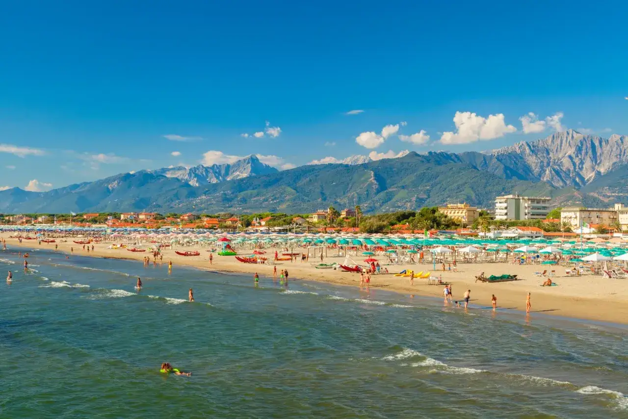 Una giornata di sole a Marina di Pietrasanta, con la spiaggia affollata di ombrelloni e bagnanti, e le montagne sullo sfondo.