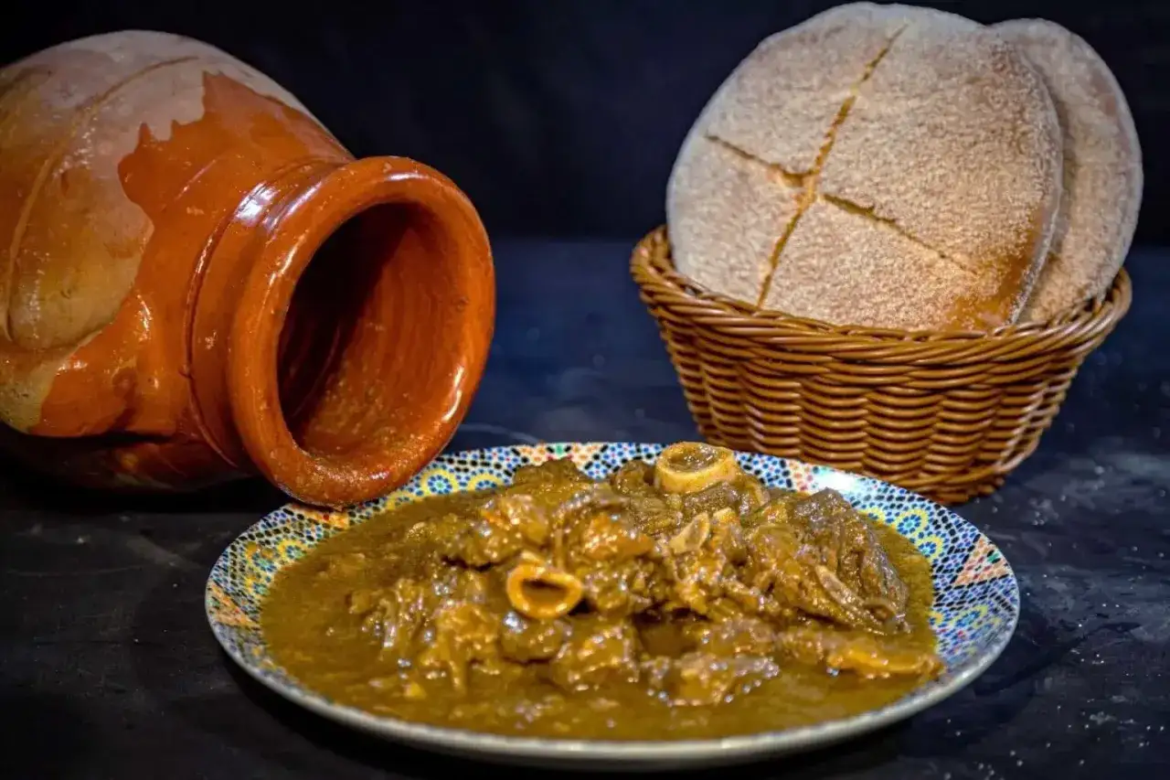 Un saporito stufato di carne in un piatto decorato, accanto a un'antica pentola di terracotta e pane fresco. Un assaggio di cosa mangiare a Marrakech.