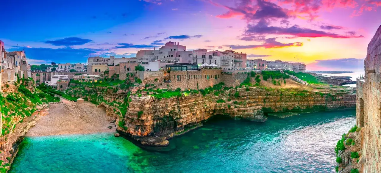 Panoramica di Polignano a Mare al tramonto: case bianche su scogliere a picco, mare turchese e cielo colorato. Perfetto per scoprire cosa fare a Polignano a Mare.