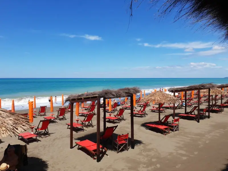Spiaggia attrezzata a Marina di Bibbona con lettini rossi, ombrelloni di paglia e il mare azzurro sullo sfondo.