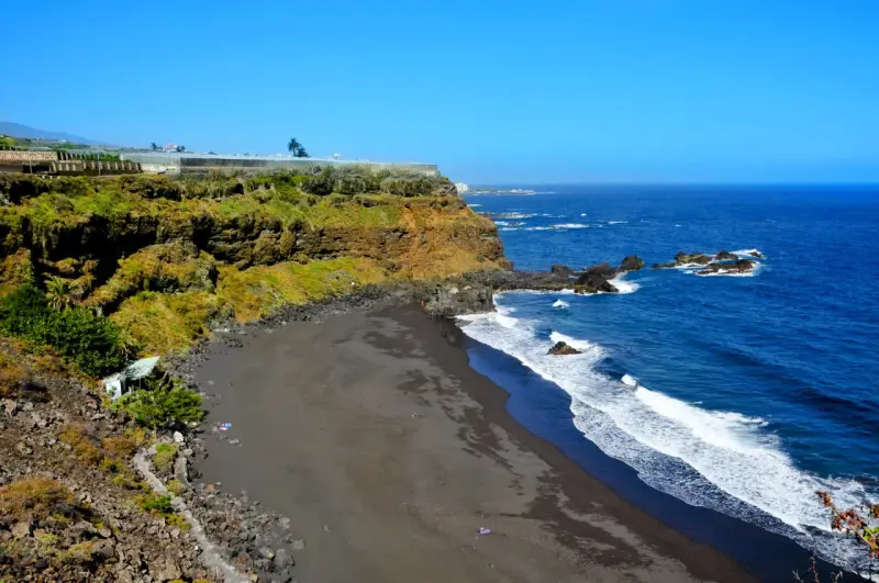 Spiaggia di sabbia nera e scogliere verdi sulle isole delle Canarie, con il blu intenso dell'oceano Atlantico.