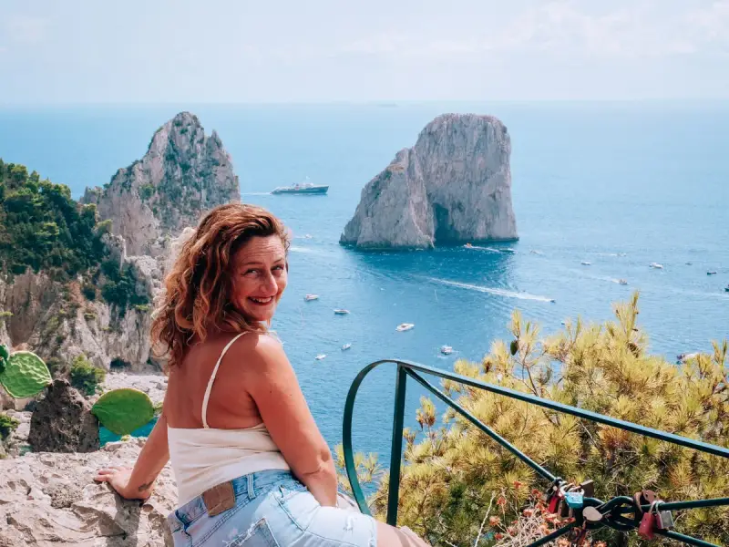 Donna sorridente con vista sui Faraglioni, un'icona dell'isola di Capri. Il mare azzurro e le barche completano il paesaggio.