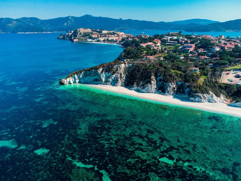 Vista aerea di una spiaggia bianca e mare cristallino, con un borgo arroccato su una collina. Un angolo di paradiso tra le isole arcipelago toscano.