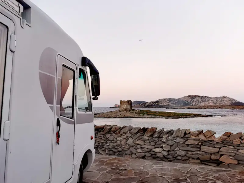 Un camper bianco parcheggiato su un lastricato di pietra, con una vista mozzafiato sul mare e una torre antica in lontananza. Un sogno di **Sardegna in camper**.